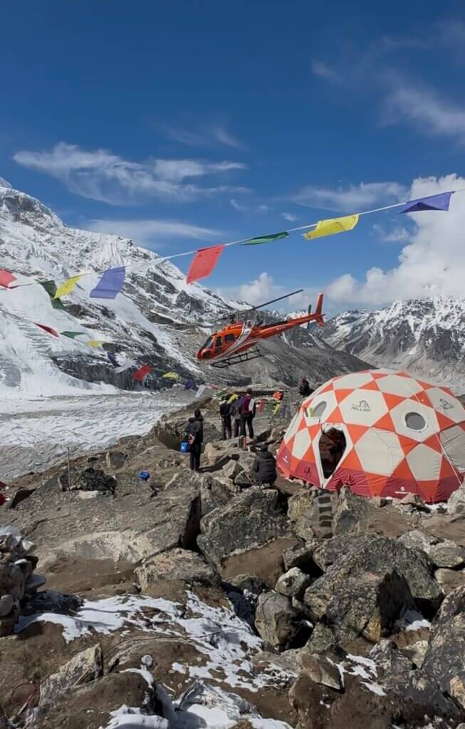 A red helicopter above a snowy mountain base camp with prayer flags and tents